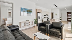 Een moderne woonkamer met een bank, fauteuils en een salontafel. Achtergrond is een keuken met een koelkast en magnetron.