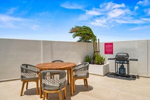 A patio with a round wooden table and four wicker chairs, a barbecue grill, and a potted plant.