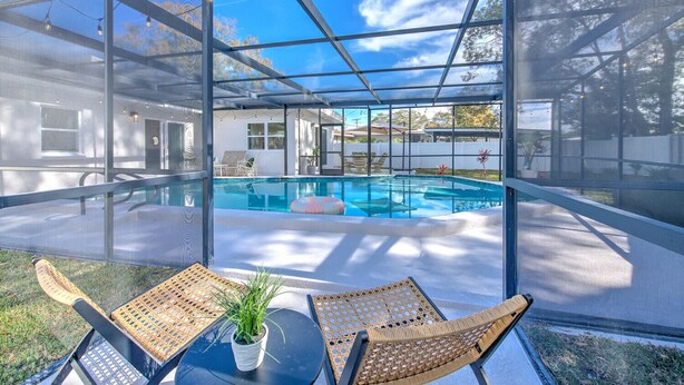 A glass-enclosed swimming pool with a view of a house and trees. There are two wicker chairs and a small potted plant on a table in the foreground. - The Peacock Place – Family Home w/Heated Pool (Tampa)
