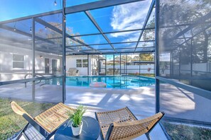 Une piscine intérieure, entourée de parois en verre, offre une vue sur une maison et des arbres. On aperçoit deux fauteuils en osier et une petite plante en pot sur une table au premier plan.