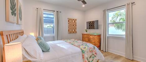 A bedroom with a bed, a wooden nightstand, a ceiling fan, a dresser, and a window with curtains.