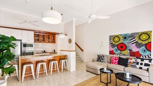 Eine moderne Küche mit weißen Schränken, einer zentralen Kochinsel mit Hockern, einem Essbereich mit einer Couch und einem farbenfrohen abstrakten Gemälde an der Wand.