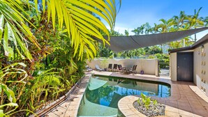 Ein Pool mit geschwungener Kante, umgeben von einer gefliesten Terrasse, Palmen und einem schattigen Bereich mit Liegestühlen.