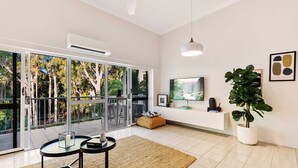 Ein modernes Wohnzimmer mit einem großen Schiebeglastür, die auf einen Balkon führt, einem wandmontierten Flachbildfernseher, einer Topfpflanze und einem kleinen runden Tisch mit einer Lampe.