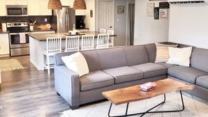 A modern living room with a grey sofa, a wooden coffee table, and a kitchen area in the background. - Our Happy Place: Hot Tub & Trail Access: STR25-00043 (Tory Hill)