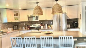 A modern kitchen with white cabinetry, a central island with chairs, and two woven pendant lights. - Our Happy Place: Hot Tub & Trail Access: STR25-00043 (Tory Hill)