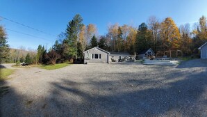 A gravel driveway leading to a wooden cabin with a garage, surrounded by trees and a clear sky. - Our Happy Place: Hot Tub & Trail Access: STR25-00043 (Tory Hill)