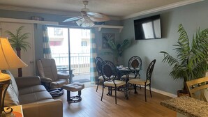 Un salon comprenant un canapé, une table à manger avec des chaises, un ventilateur de plafond et un téléviseur fixé au mur.