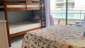 Une chambre avec un lit superposé, un lit recouvert d’une couvre-lit à motifs floraux, une fenêtre donnant sur un balcon et une pancarte indiquant : « On vous accueille les pieds nus ».