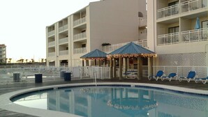 Une piscine avec une terrasse en bois, un espace de détente couvert avec des parasols et un immeuble à plusieurs étages doté de balcons.