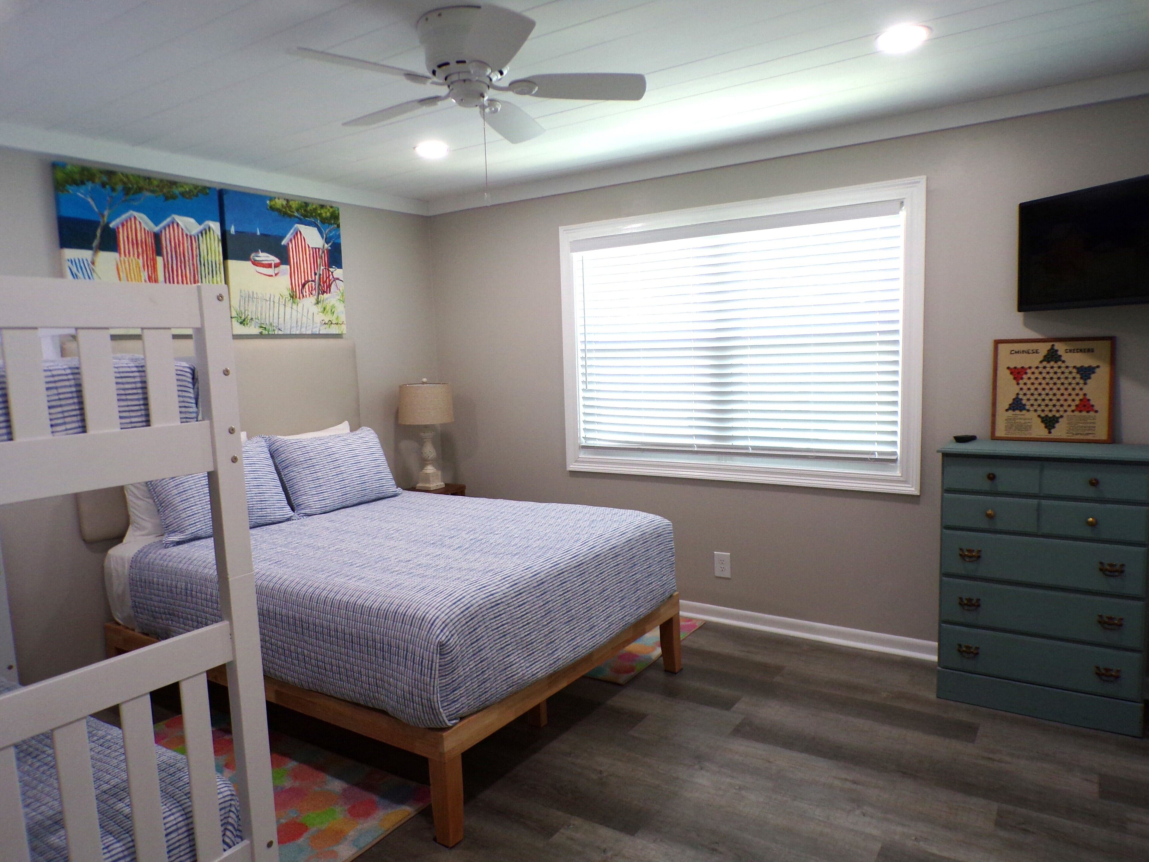 A bedroom with a bunk bed, a single bed, a ceiling fan, a window with blinds, a dresser, and a wall-mounted TV.