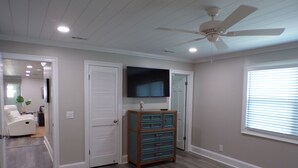 Ein Schlafzimmer mit Deckenventilator, einem an der Wand montierten Fernseher, einem Schminktisch und einem Fenster mit Jalousien.