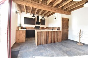 Une cuisine rustique avec des poutres en bois, un îlot central, des luminaires suspendus et un sol carrelé.