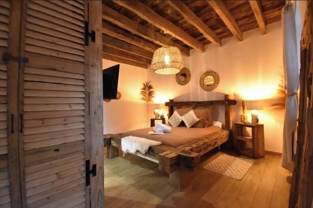 A rustic bedroom with a wooden bed frame, a hanging light fixture, and a small bedside table.