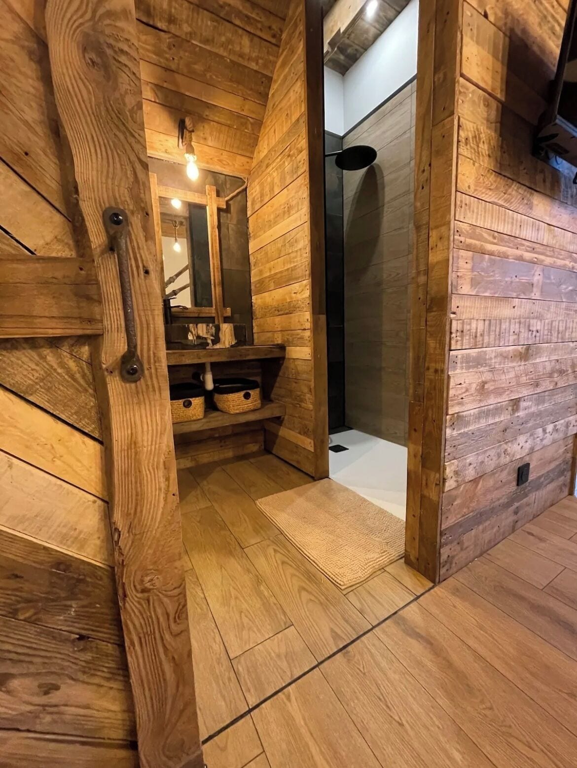 Un interior de madera con una puerta de madera, un espejo, un estante con cestas y una zona de ducha.