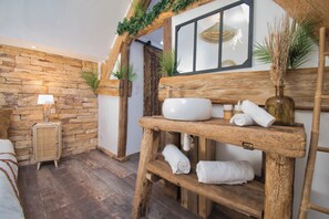 Un baño rústico con pared de piedra, vigas de madera, un estante de madera con toallas, un lavabo blanco y una mesita de noche con una lámpara.