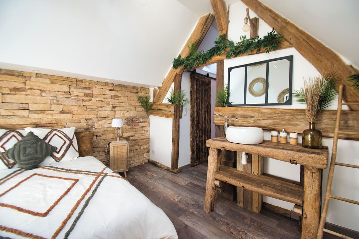 A bedroom with a stone wall, wooden beams, a bed with patterned bedding, a bedside table with a lamp, a wooden dresser with a mirror, and a rustic ceiling.