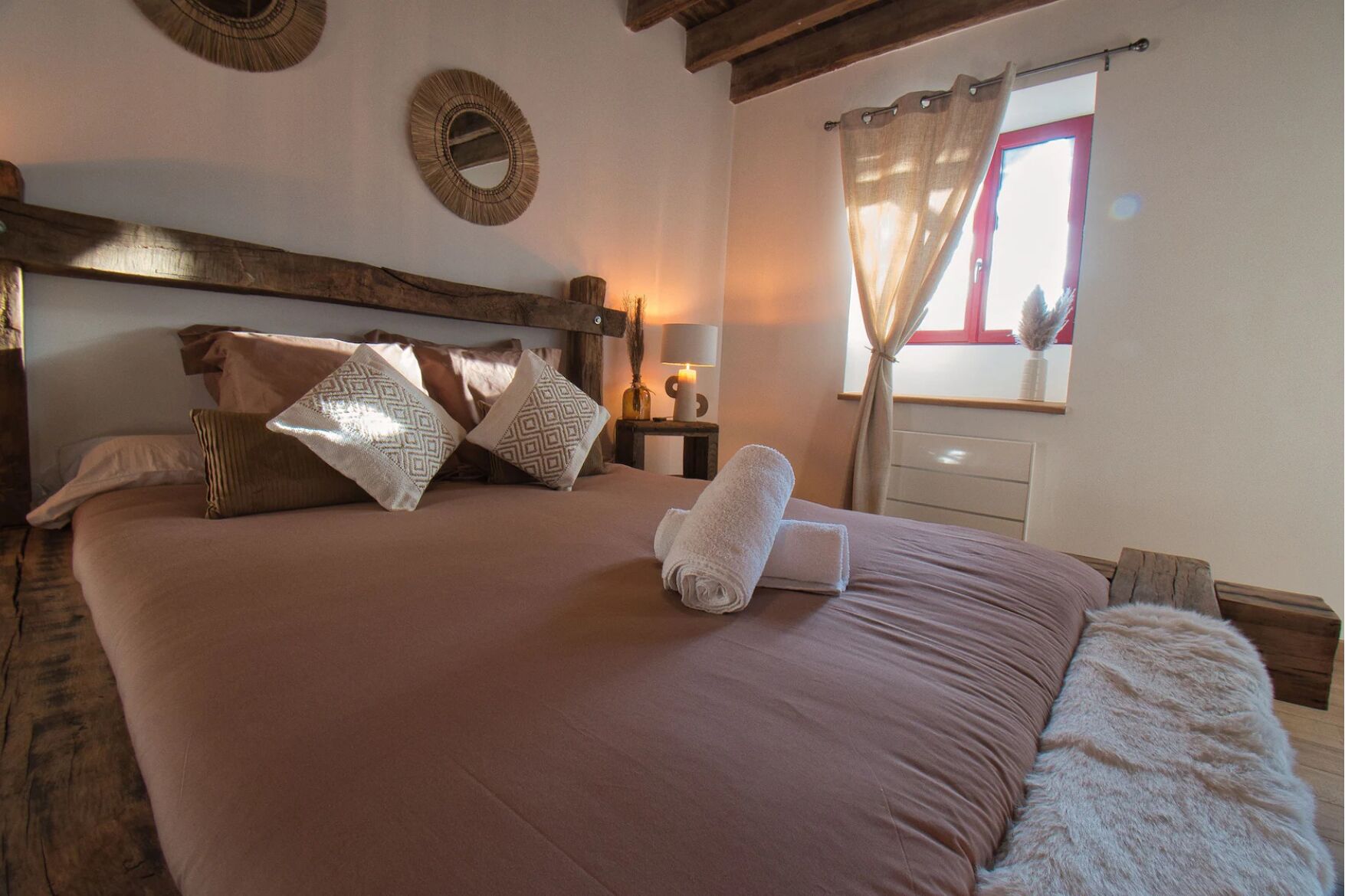 A large bed with a wooden headboard, multiple pillows, a bedside lamp, and a window with curtains.