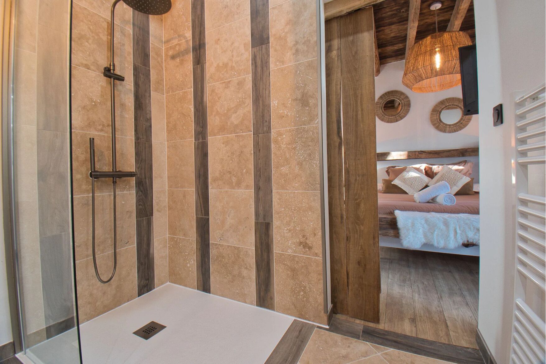 A modern bathroom with a shower, a wooden ceiling, and a bed visible through an open door.