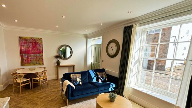 A living room with a dark blue sofa, a round wooden coffee table, a dining table with chairs, a large abstract painting, a round mirror, and a window with curtains. - Charles Hope Salisbury Centre (Salisbury)