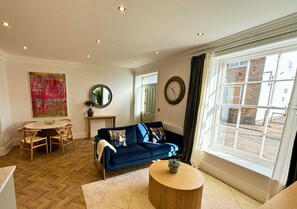 A living room with a dark blue sofa, a round wooden coffee table, a dining table with chairs, a large abstract painting, a round mirror, and a window with curtains. - Charles Hope Salisbury Centre (Salisbury)