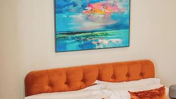 Une chambre avec un grand lit, du linge de lit blanc, deux oreillers orange, une table de chevet avec une lampe et un tableau abstrait coloré accroché au mur.