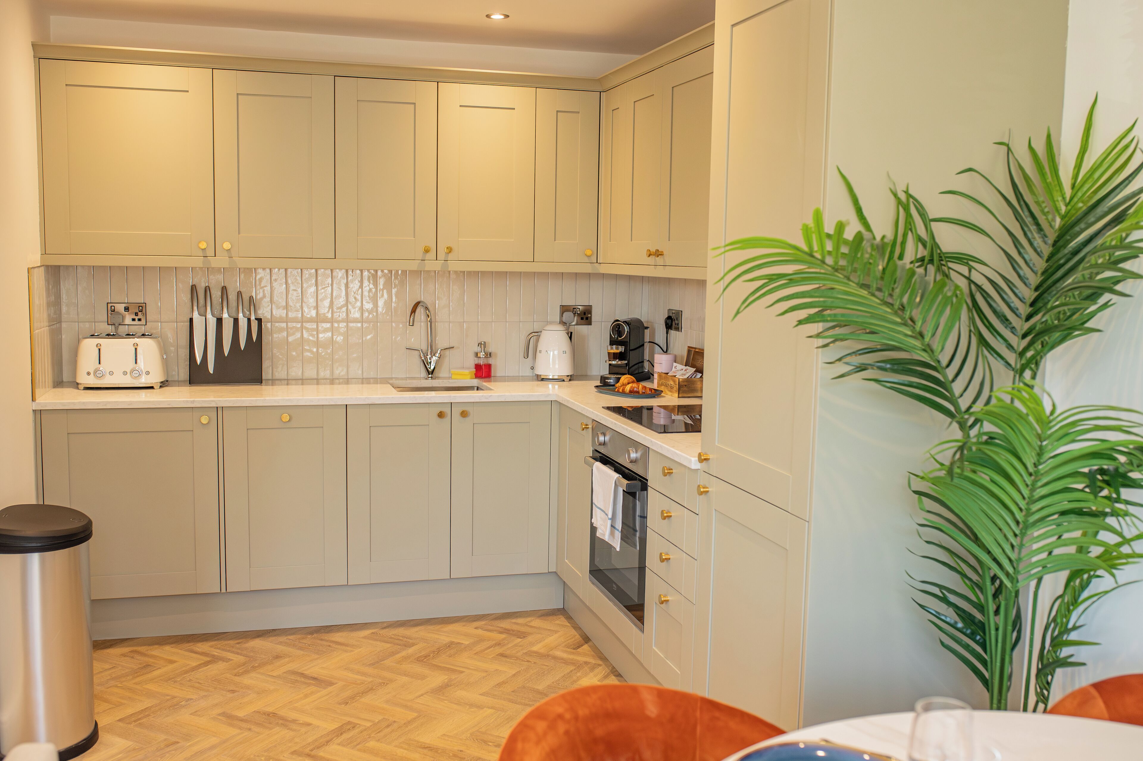 A kitchen with light-colored cabinets, a tiled backsplash, a sink, a toaster, a kettle, and a coffee maker. There is a trash can and a potted plant.