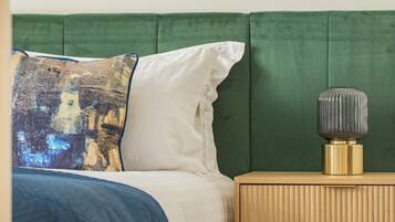 Un dormitorio con un cabecero tapizado en verde, una almohada blanca, una manta azul y una mesita de noche con una lámpara.