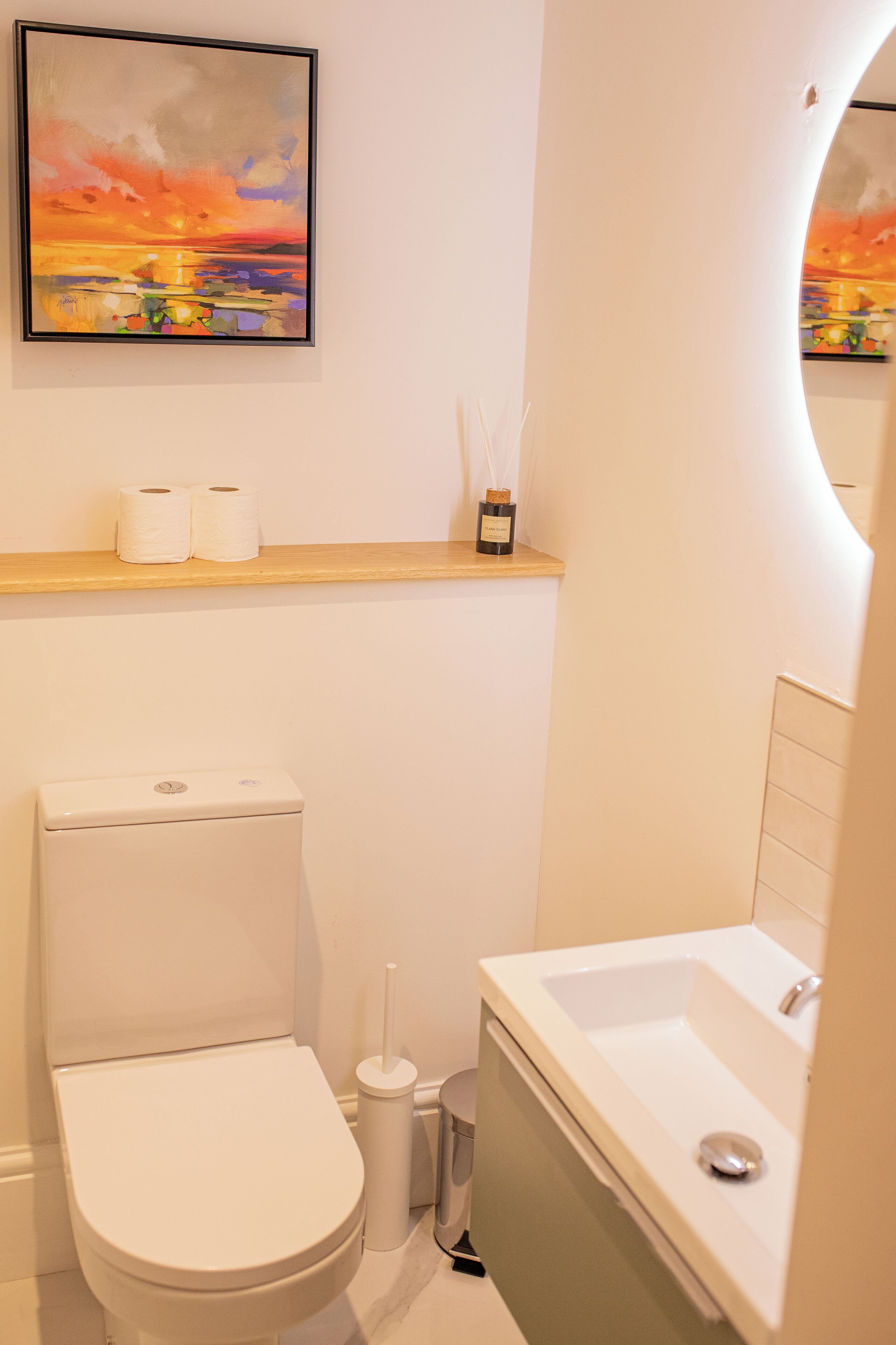 Un baño moderno con inodoro, lavamanos y una repisa con papel higiénico y una vela.