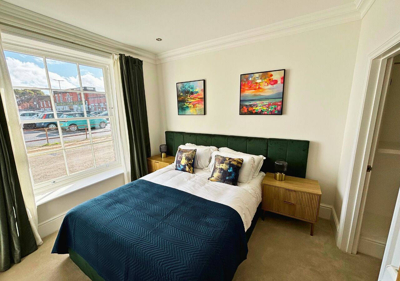 A hotel room with a large bed, a green headboard, two paintings on the wall, a window with a view of a parking lot, and a bedside table with a lamp.