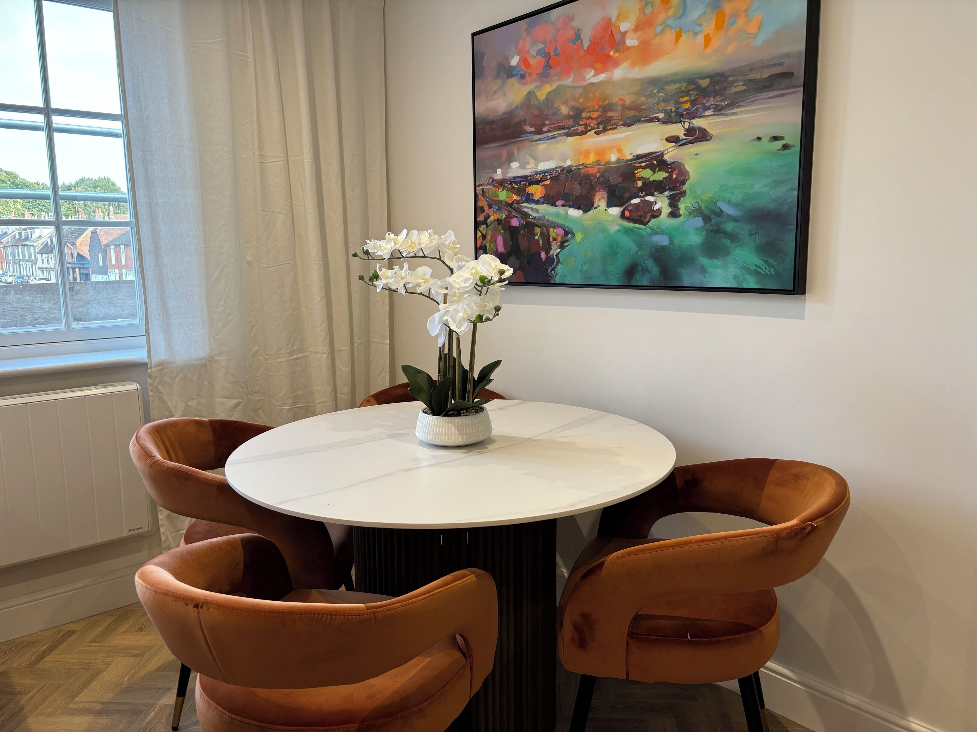 Een eetgedeelte met een ronde tafel, vier stoelen, een potorkidee en een groot abstract schilderij aan de muur.