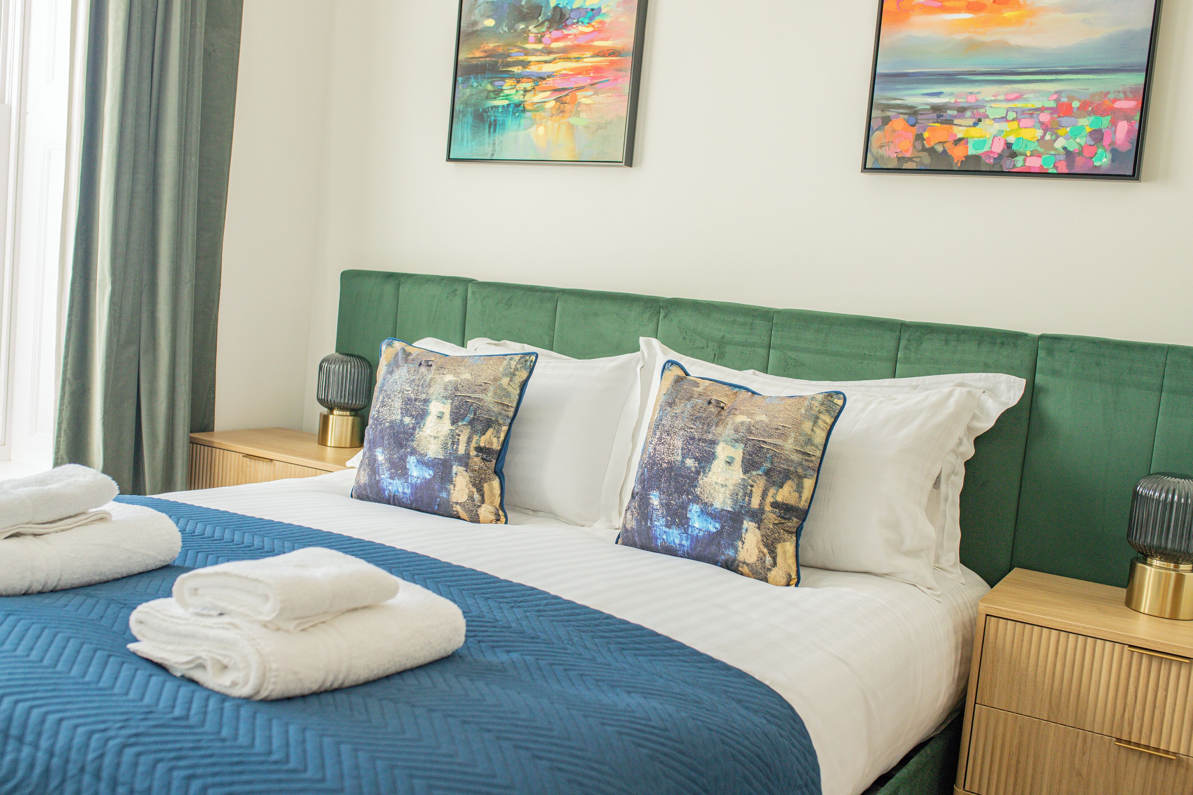 A neatly made bed with a blue and white bedspread, green headboard, and two decorative pillows. There are two bedside tables with lamps and folded towels on the bed.
