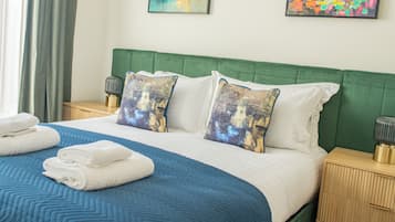 A neatly made bed with a blue and white bedspread, green headboard, and two decorative pillows. There are two bedside tables with lamps and folded towels on the bed.