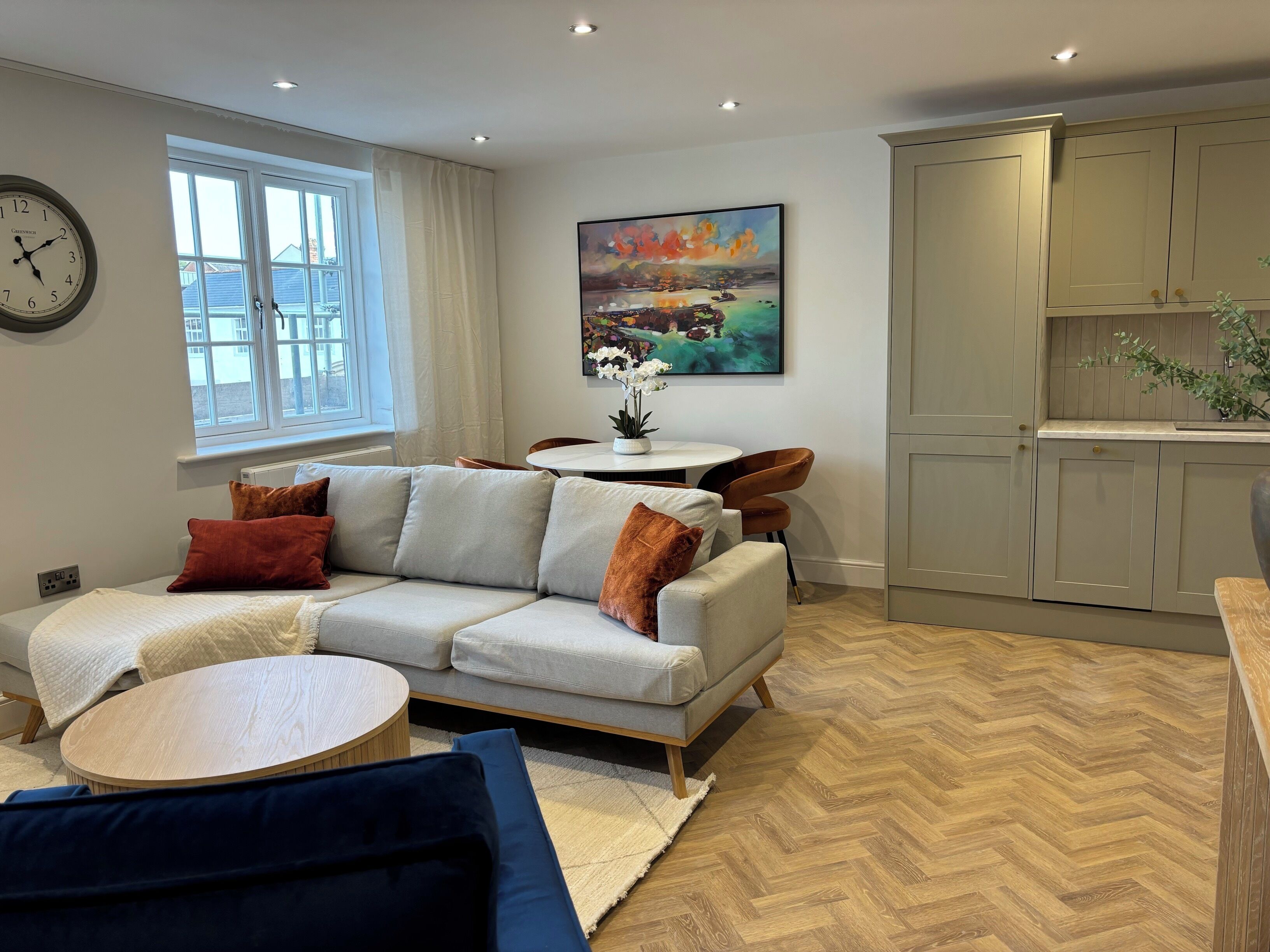 A modern living room with a sofa, a round coffee table, and a dining table with chairs. There is a painting on the wall, a window with curtains, and a kitchen area with cabinets.
