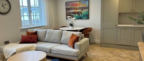 A modern living room with a sofa, a round coffee table, and a dining table with chairs. There is a painting on the wall, a window with curtains, and a kitchen area with cabinets.