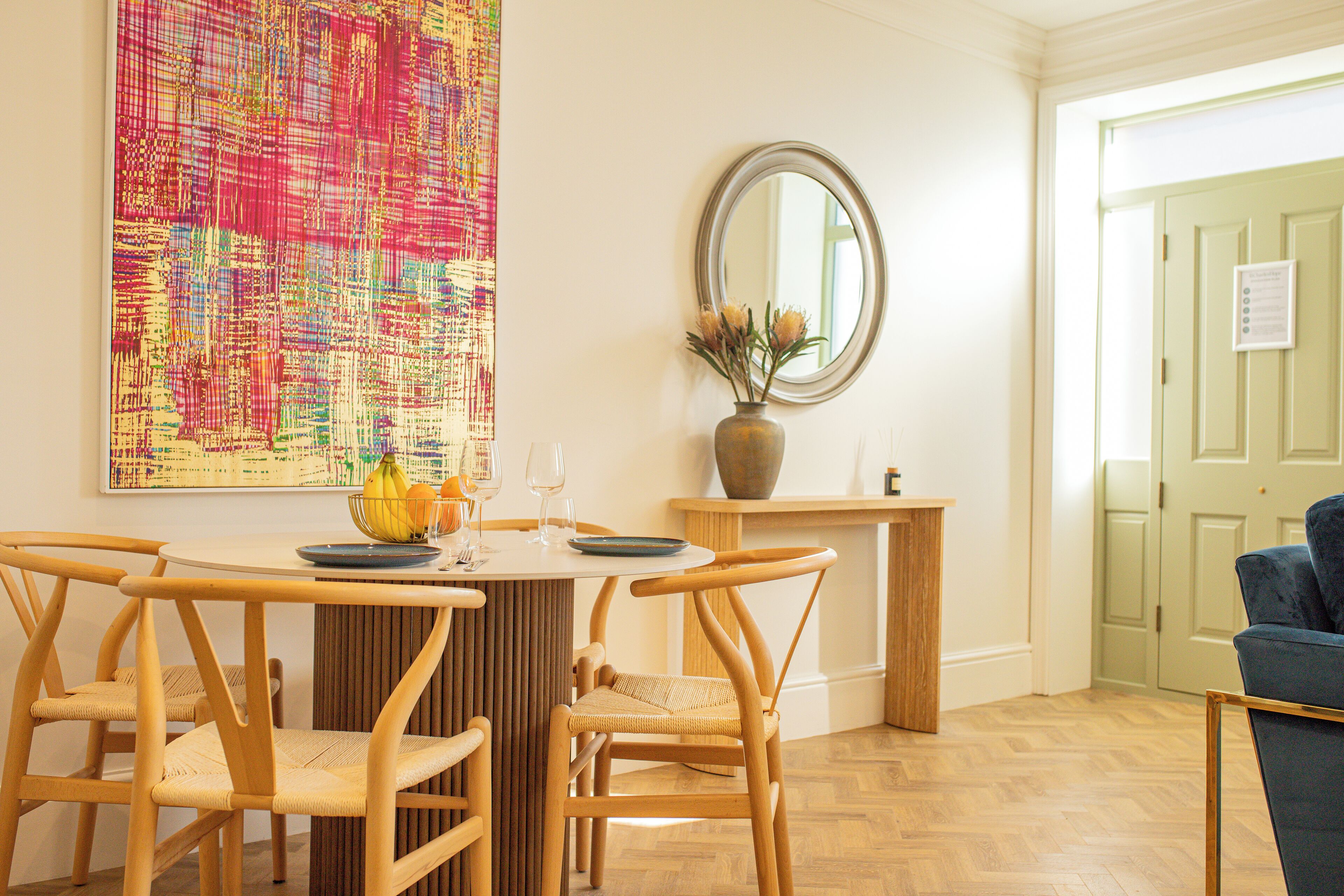 A dining area with a round table, four wooden chairs, a vase with flowers, and a large abstract painting on the wall.