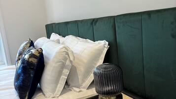 A neatly made bed with white and gold pillows, a green upholstered headboard, and a bedside table with a lamp and a drawer.