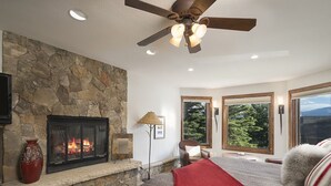 Une chambre avec une cheminée en pierre, un ventilateur de plafond, un lit recouvert d’une couverture rouge et de grandes fenêtres donnant sur la forêt.