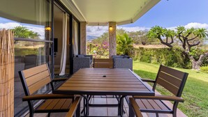 A patio with a wooden table and chairs, a glass door leading outside, and a view of a garden with trees and shrubs. - Kamaole Beach 1 Condo at Kihei Ali'i Kai (Kihei)