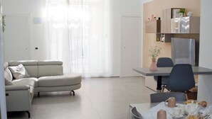 En moderne stue med en beige sofa, et spisebord med stole og et køkkenområde med et køleskab og reoler.