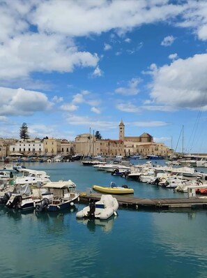 Un port de plaisance avec des bateaux amarrés, un bâtiment imposant surmonté d’une tour, et un ciel dégagé parsemé de nuages.