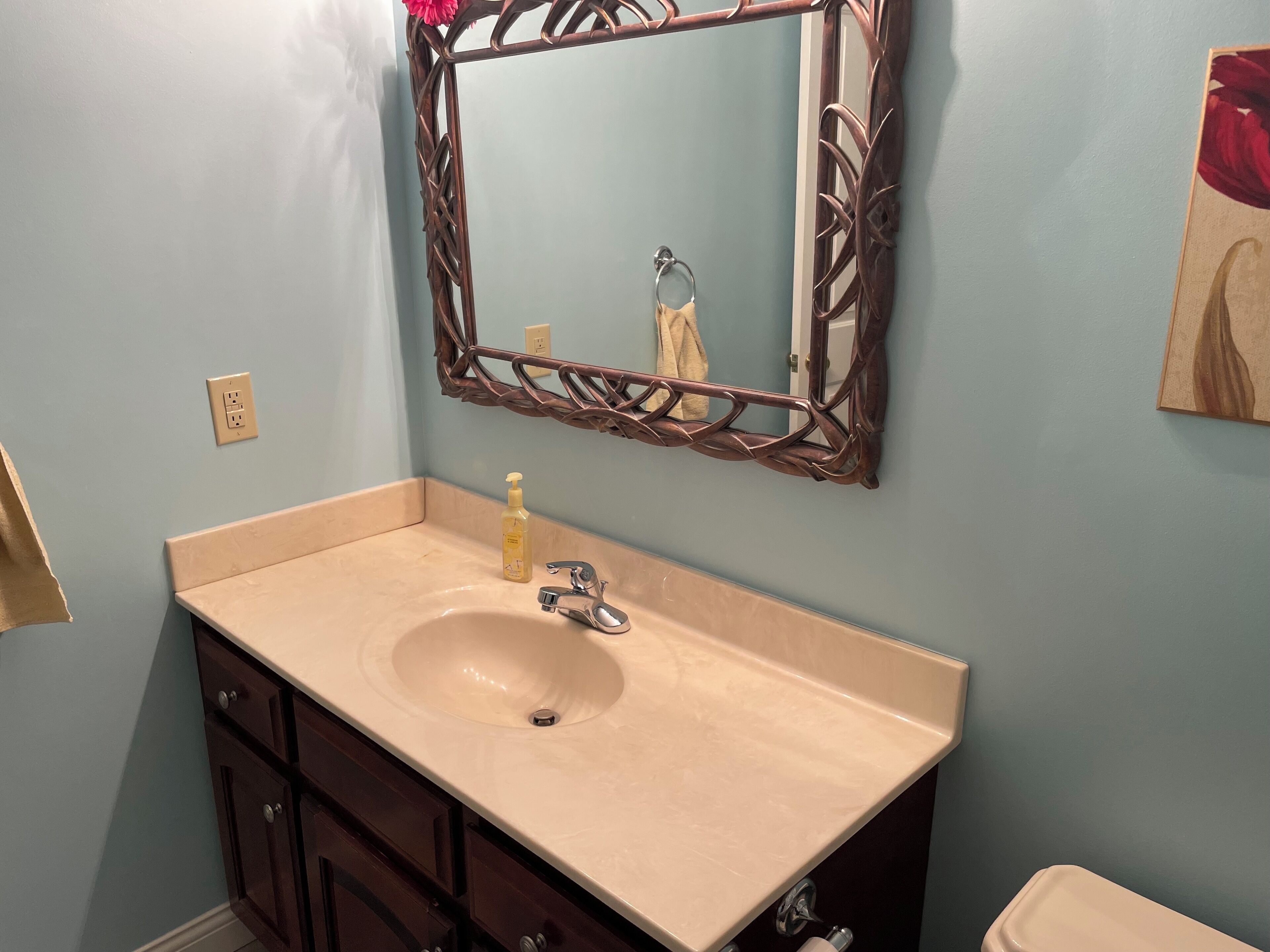 A bathroom with a beige sink, dark wooden cabinets, a mirror with a decorative frame, and a towel hanging on the wall.