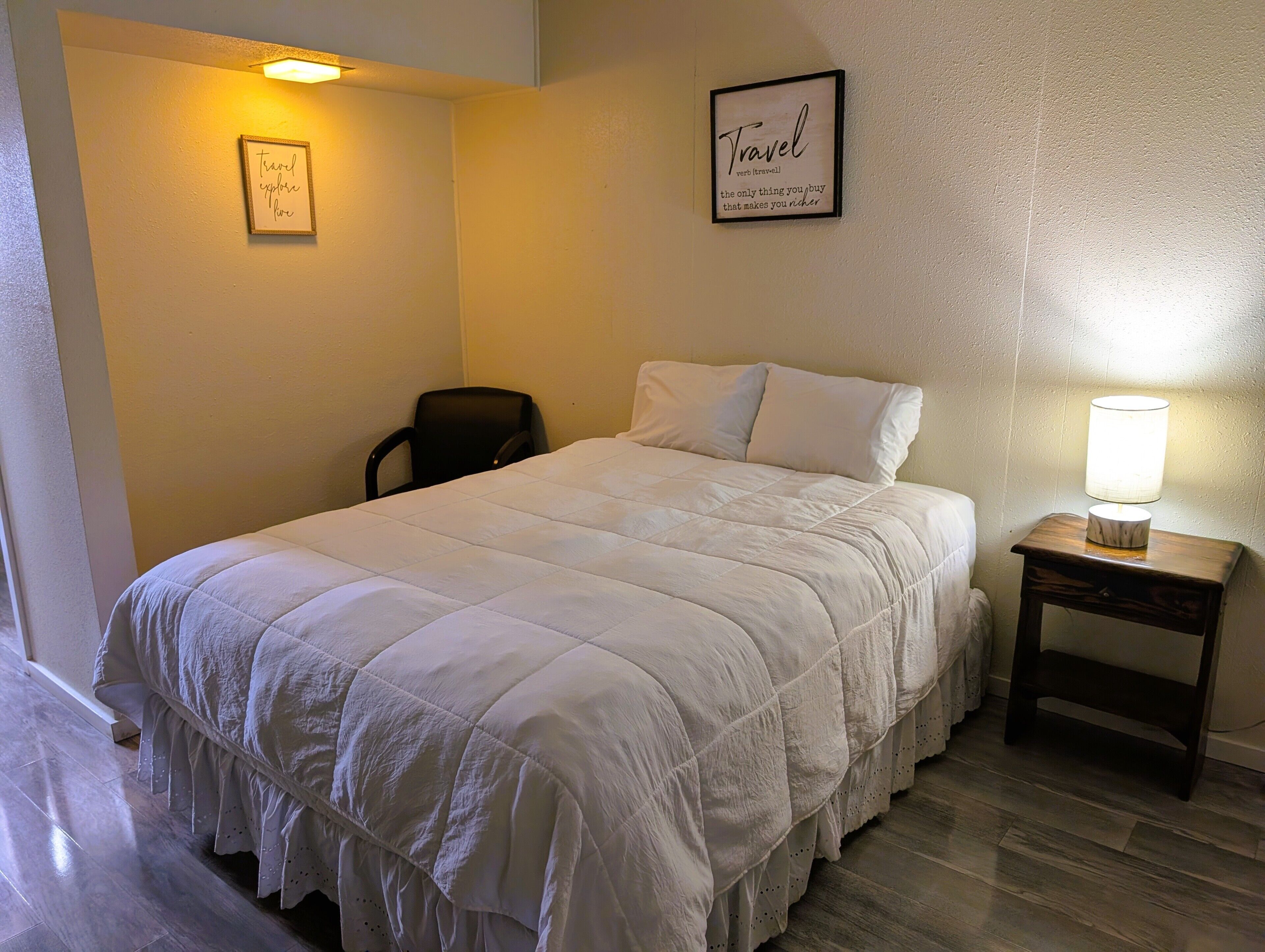 Un lit bien fait, recouvert d’une couette blanche, une petite table de chevet avec une lampe, une chaise et deux citations encadrées accrochées au mur.