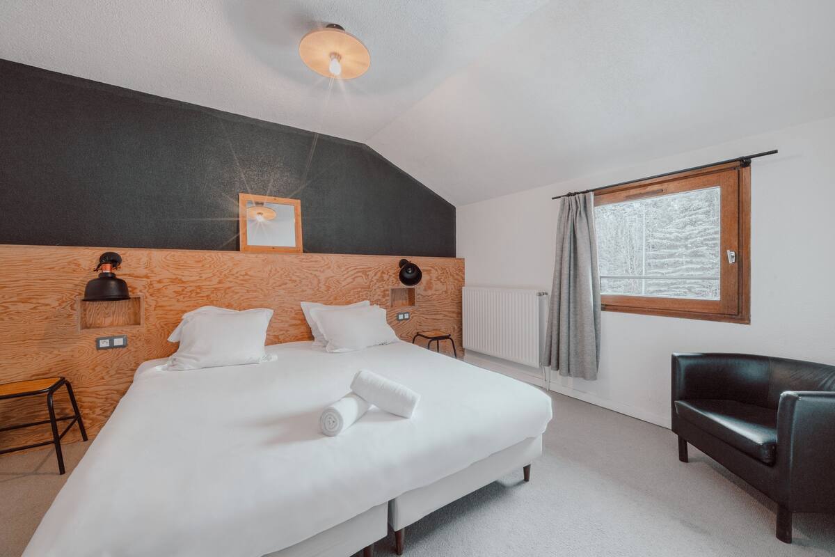 A bedroom with a large bed, a wooden headboard, a window with curtains, a black leather chair, and a wall-mounted lamp.