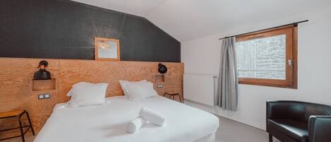 A bedroom with a large bed, a wooden headboard, a window with curtains, a black leather chair, and a wall-mounted lamp.