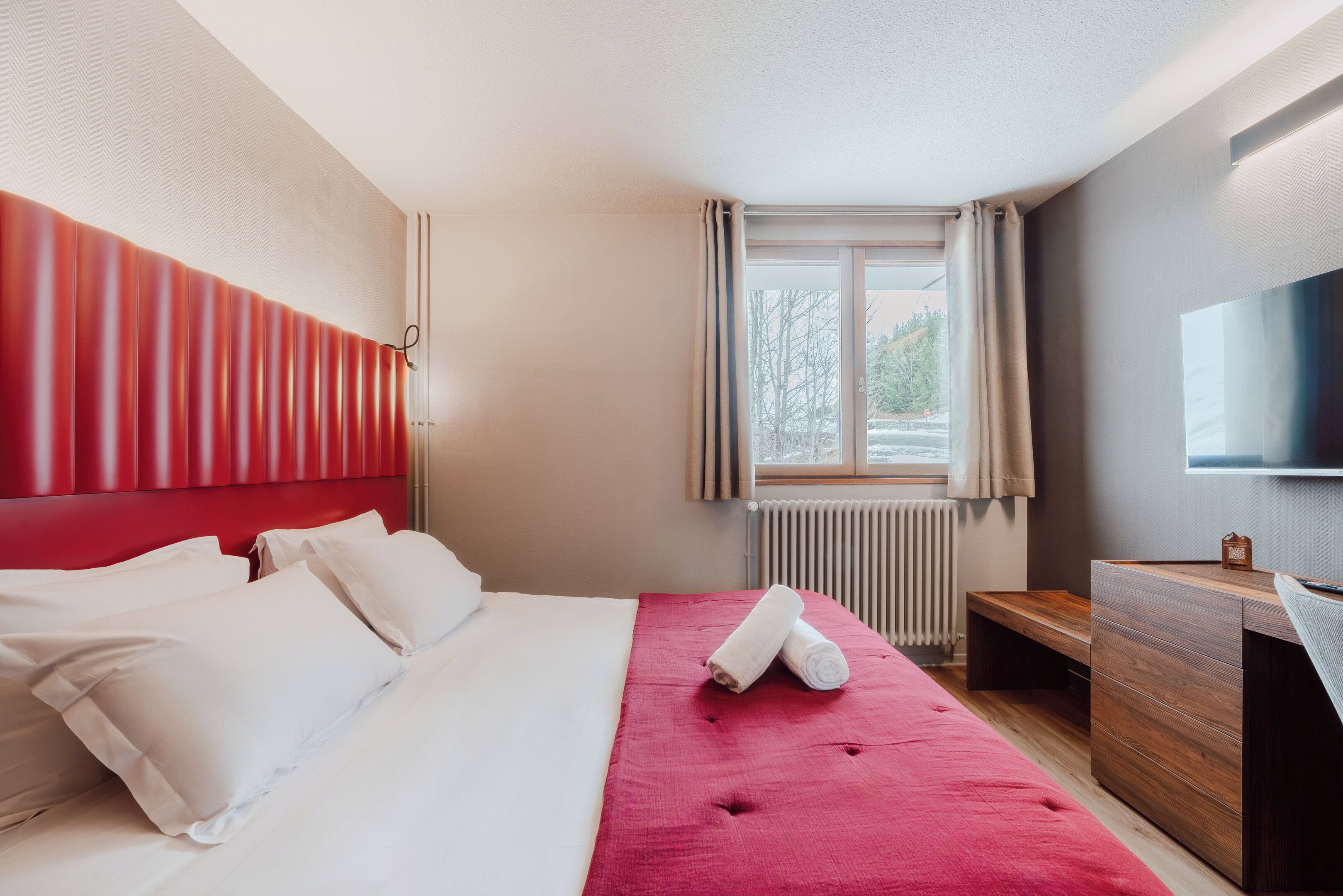 A hotel room with a bed, a red headboard, a window with curtains, a wooden desk, and a flat-screen TV.