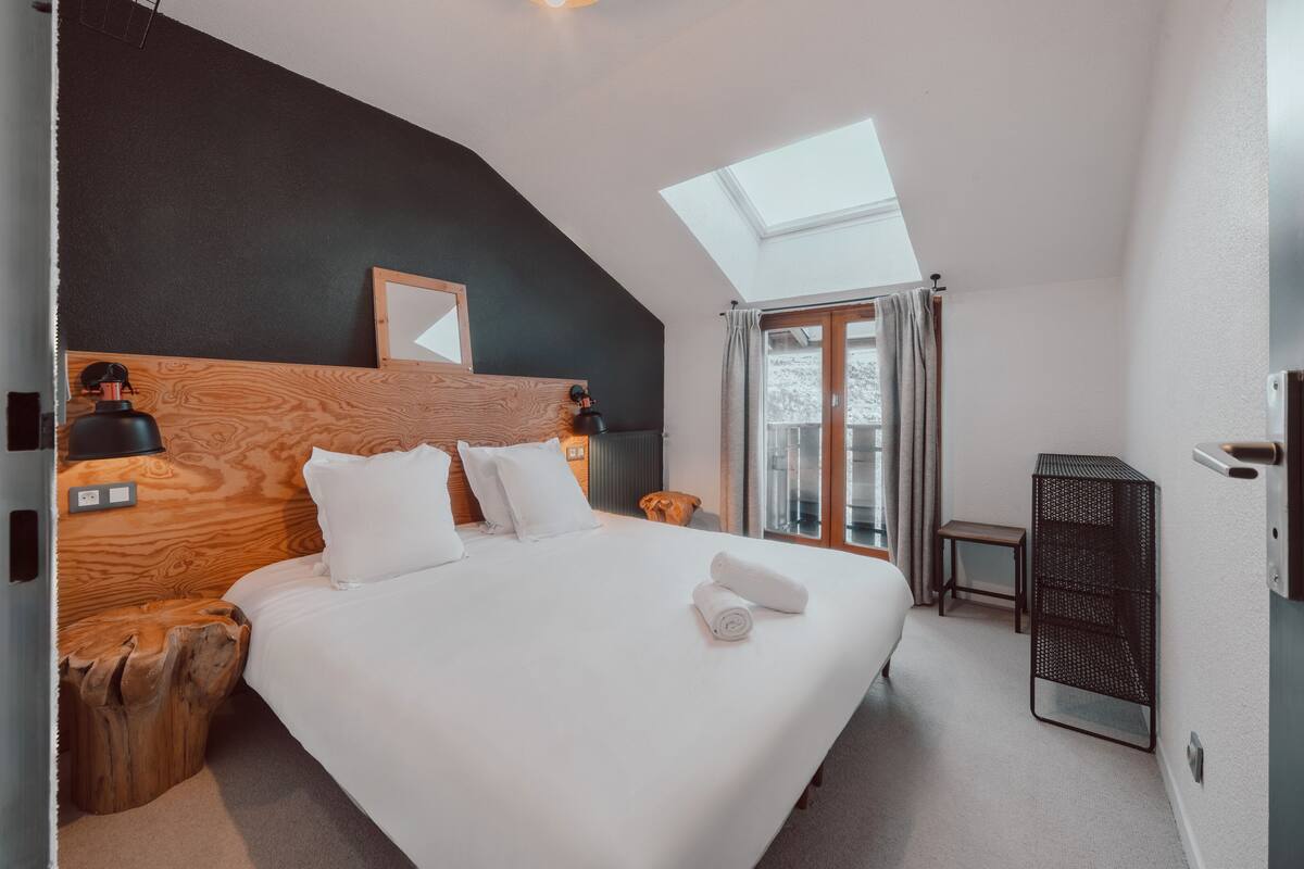 A modern hotel room with a large bed, wooden headboard, skylight, and a small side table.