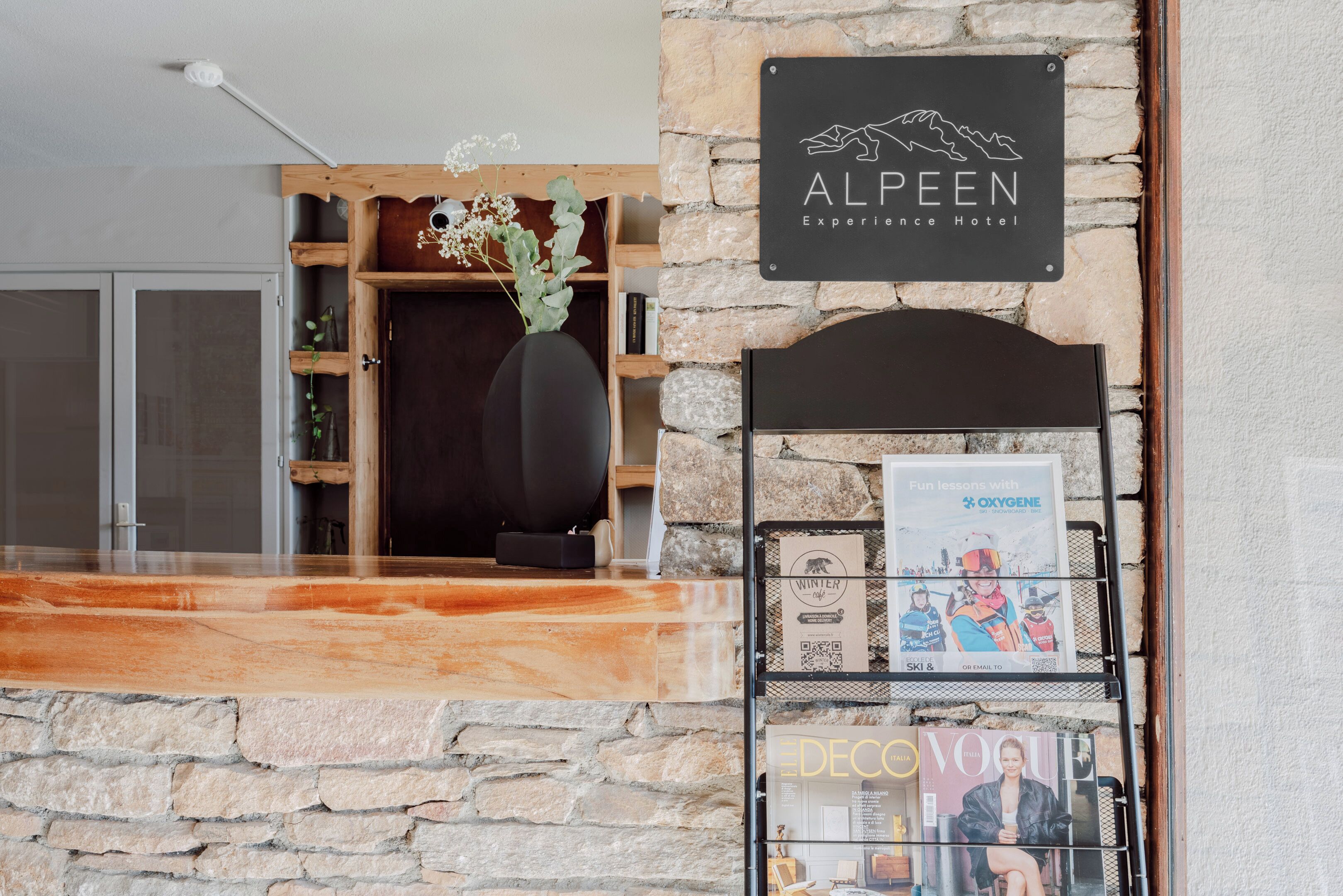 A reception area with a stone wall, a wooden reception desk, a sign for 'ALPEEN Experience Hotel', and a magazine rack with 'Vogue' and 'OXYGENE' magazines.