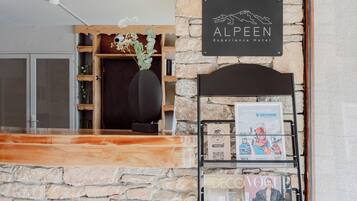 A reception area with a stone wall, a wooden reception desk, a sign for 'ALPEEN Experience Hotel', and a magazine rack with 'Vogue' and 'OXYGENE' magazines.