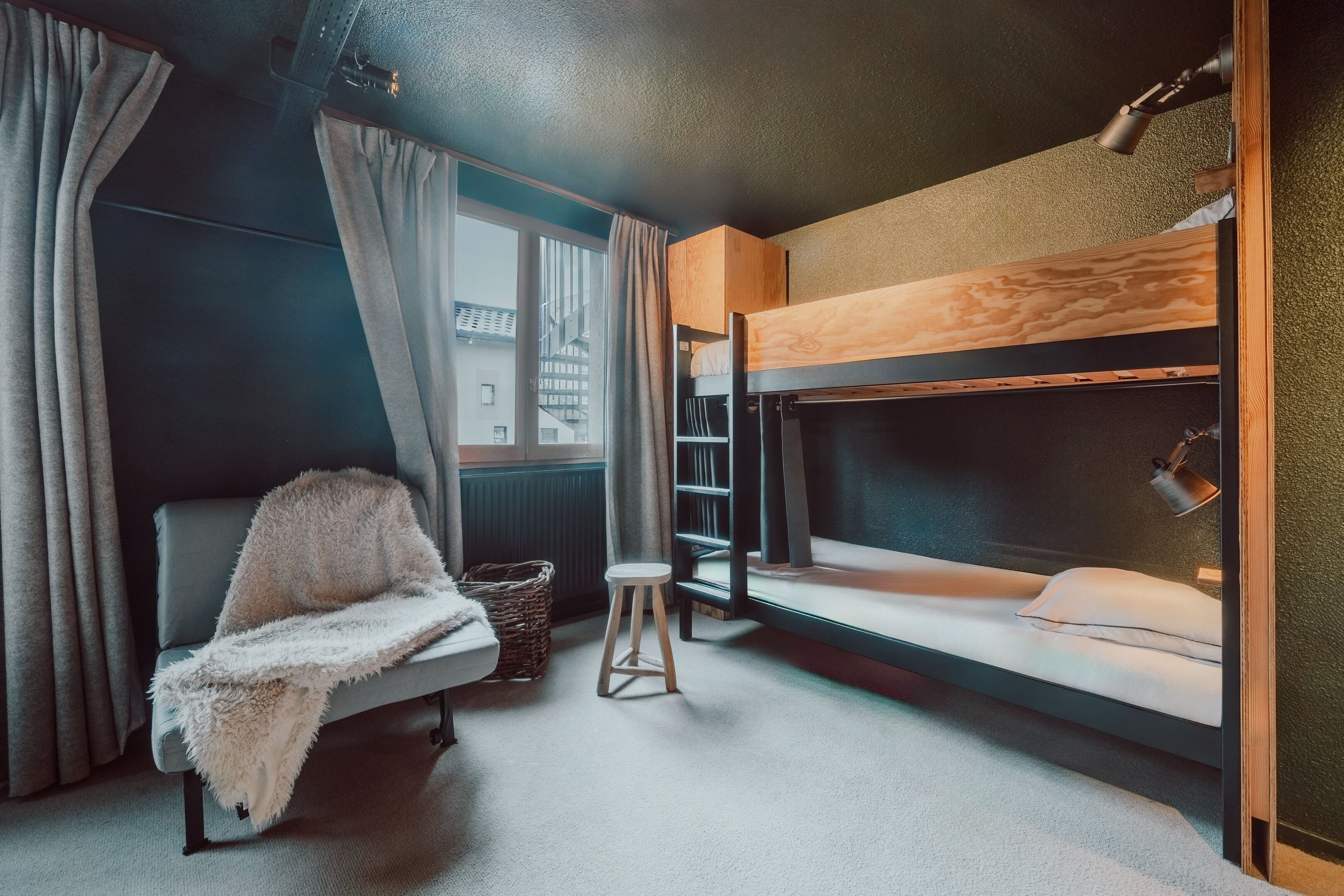 Une pièce avec des lits superposés, une chaise avec une couverture, un petit tabouret et une fenêtre avec des rideaux.